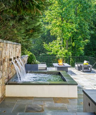 An example of small backyard ideas showing a brick wall with a water feature attached in front of a seating area with a firepit