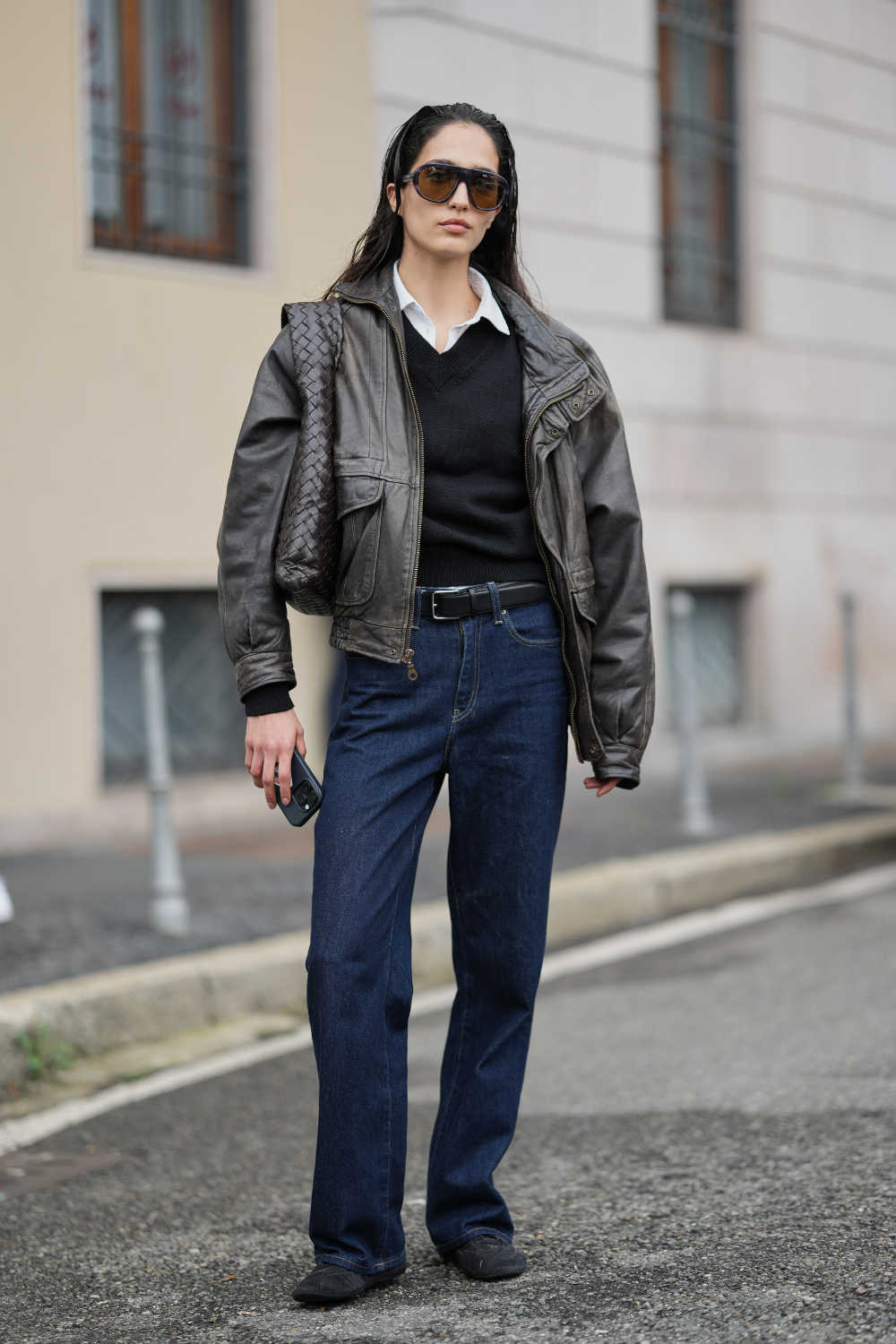 A guest wears long dark brown hair worn slicked back with a center part, oversized tortoiseshell gradient sunglasses, woven dark brown leather shoulder bag, white cotton collared shirt under a black knit v-neck sweater, dark brown leather zip-front jacket with flap pockets, black leather belt with silver buckle, dark indigo denim jeans in a straight-leg cut, black leather flat shoes, outside Tod's, during Milan Fashion Wee