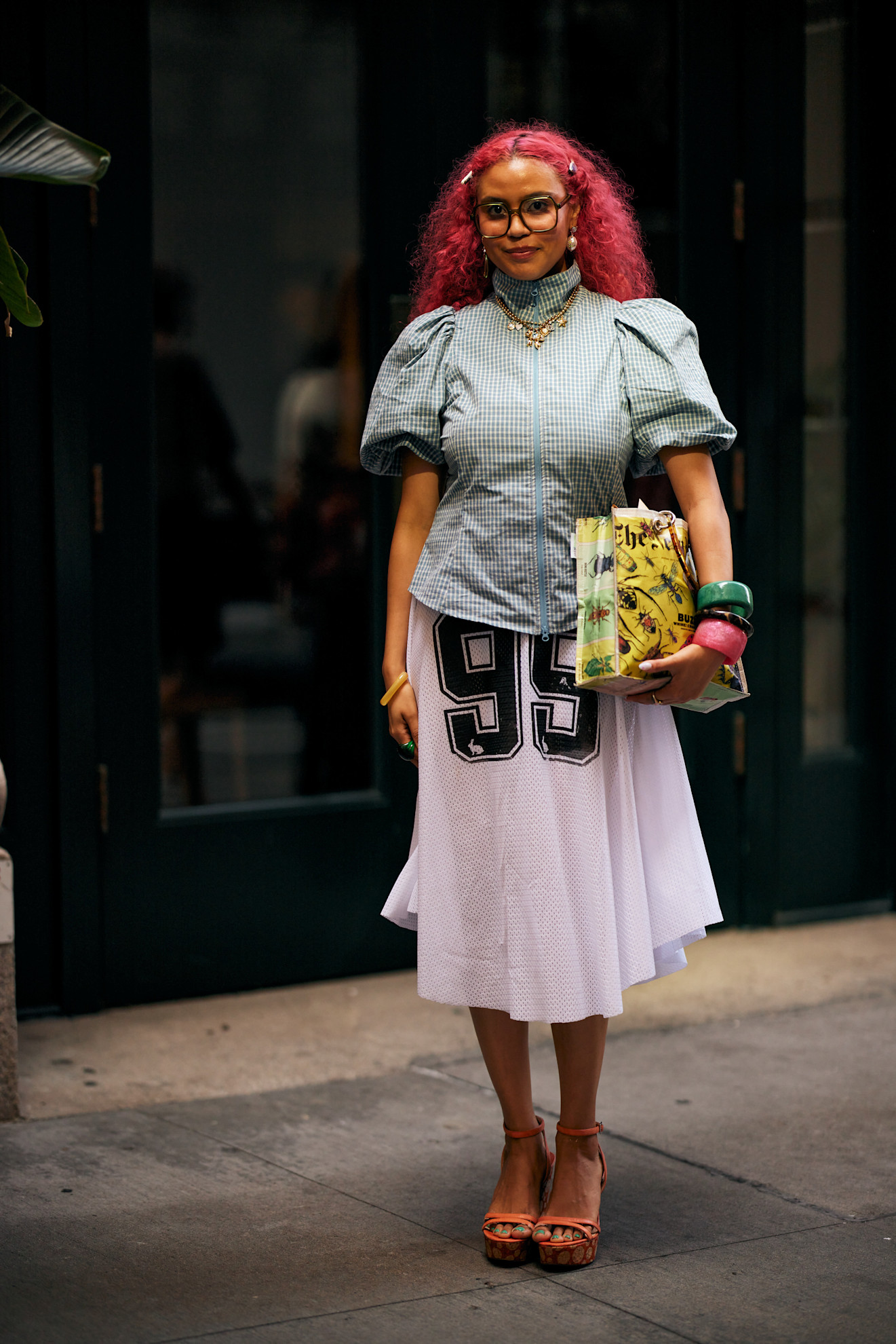 a woman with hot pink hair wearing glasses, a white skirt, and blue top with orange heels