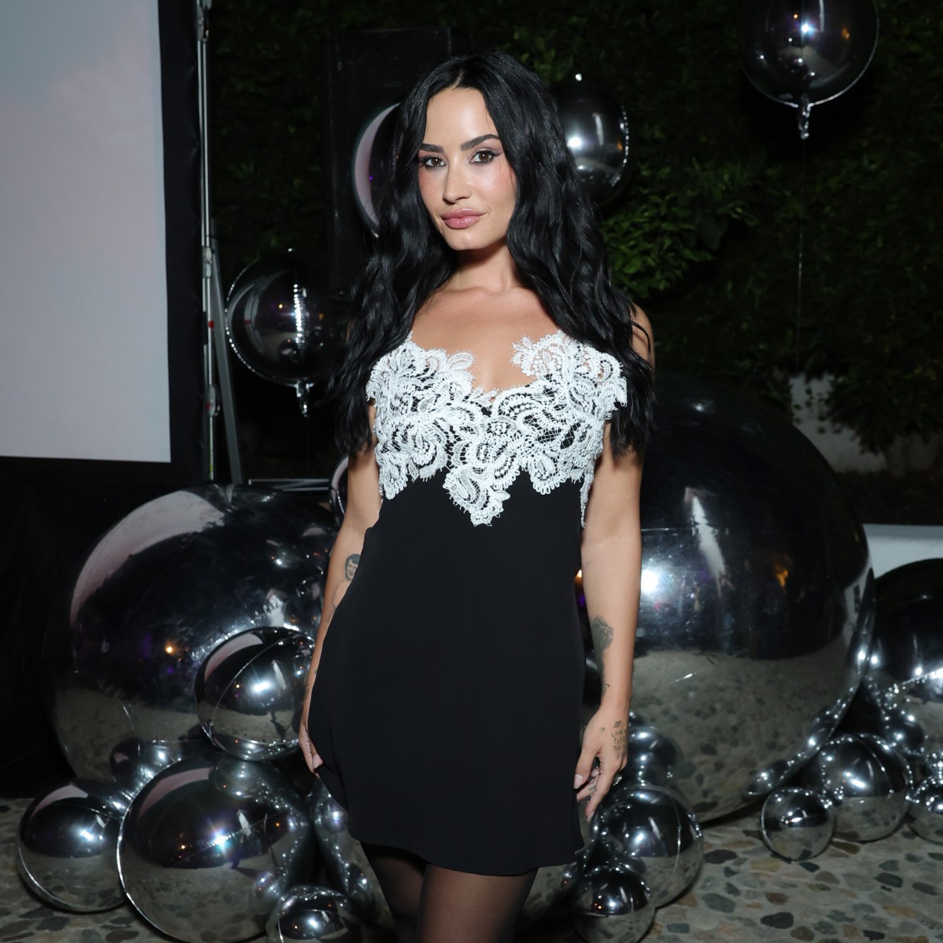 a woman with long, black hair wearing a black and white mini dress posing in front of silver balloons