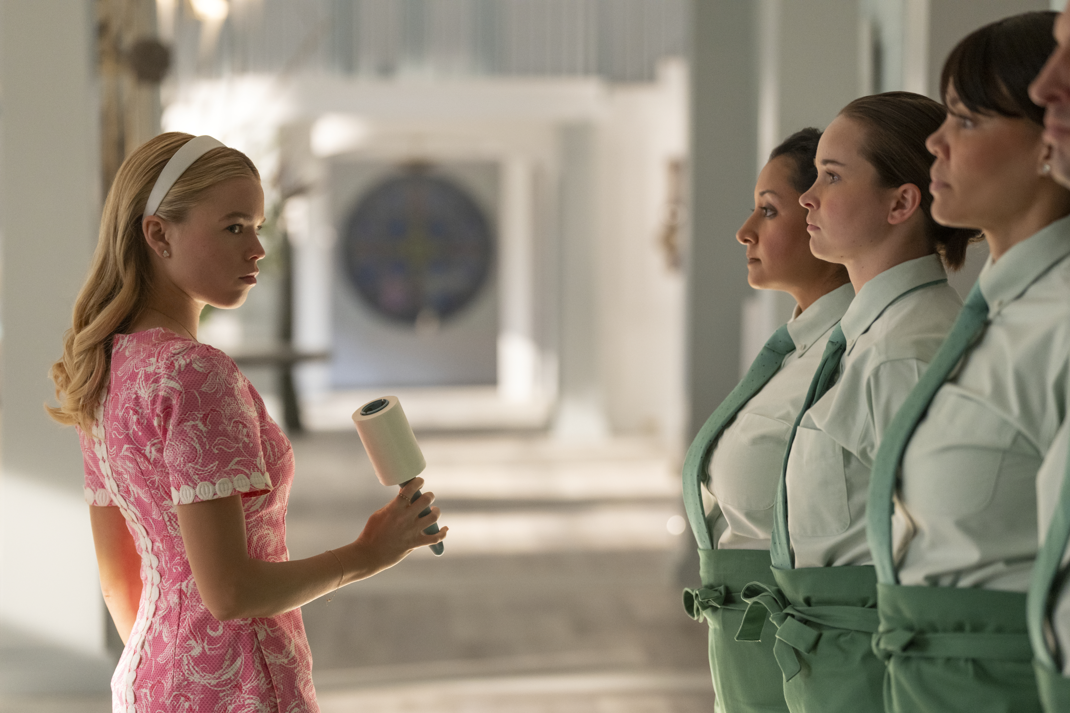 A woman (Milly Alcock as Simone) holds a lint roller as she stands in front of a line of waiters, in episode 101 of Sirens.