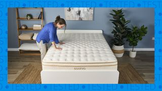 The image shows a dark haired mattress tested placing her hands on the surface of a cream and gold mattress