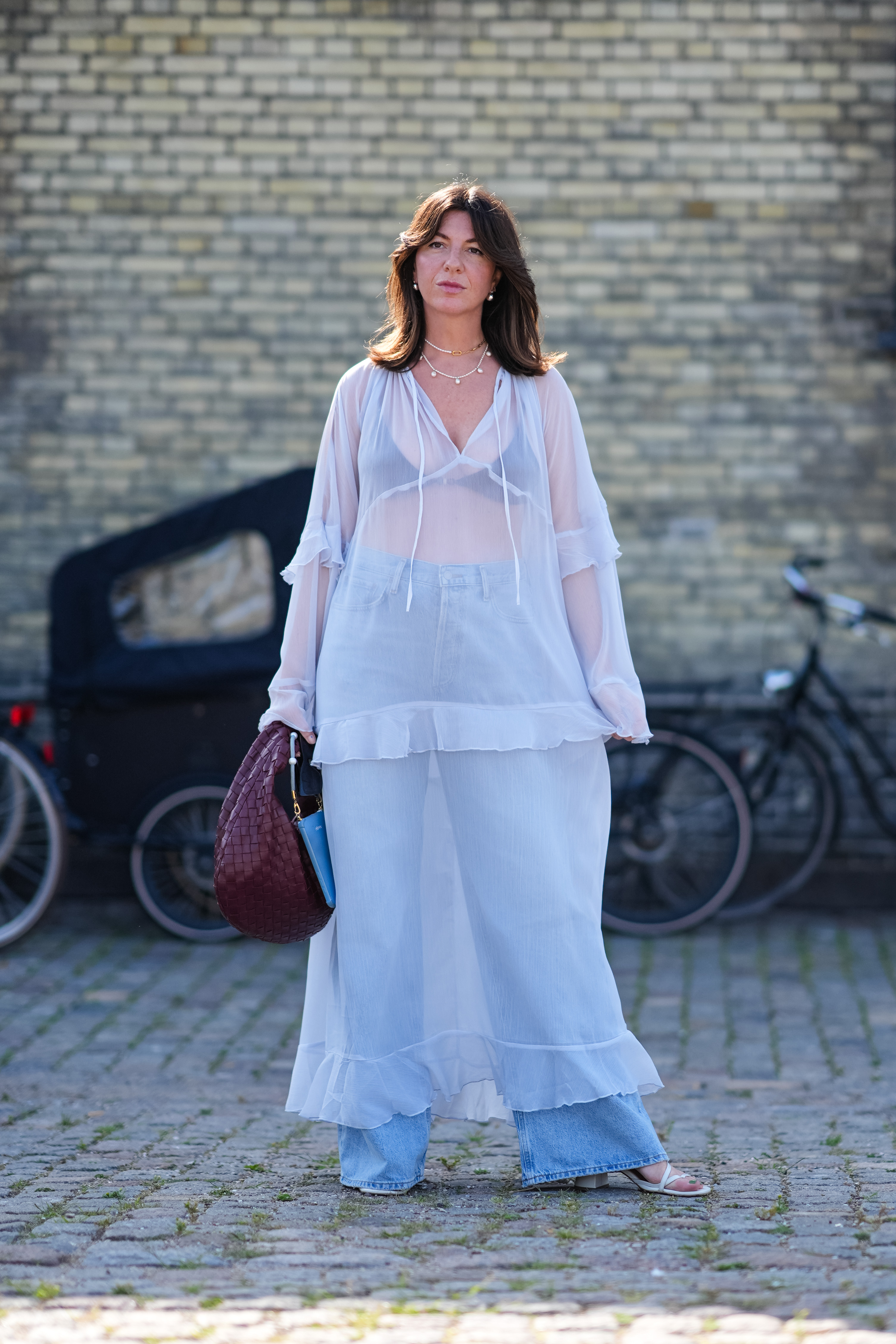 COPENHAGEN, DENMARK - AUGUST 7: A guest wears silver earrings, silver layered necklace, black bralette, light blue high waisted denim jean pants, white sheer mesh long sleeve midi dress, dark brown red woven Bottega Veneta leather bag, light blue bag, white trappy open toed heels, outside Herskind, during the Copenhagen Fashion Week Spring/Summer 2024-2025 on August 7, 2024 in Copenhagen, Denmark. (Photo by Edward Berthelot/Getty Images)