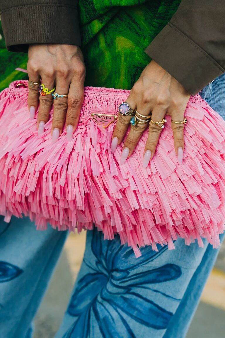 a woman's hands with long pink nails, lots of gold rings, holding a pink fringe bag and wearing blue jeans