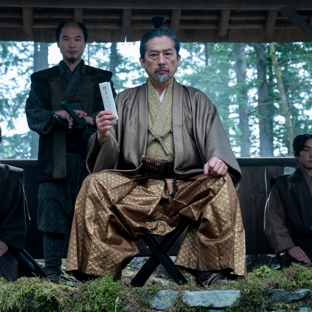 a man in gold japanese period wear (Hiroyuki Sanada as Yoshii Toranaga) sits while holding up a white letter, with one man standing behind him and two more kneeling at each side, in 'shogun' (2024)