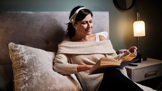 A woman lies in bed reading, a Neurovalens Modius Sleep device worn around her head