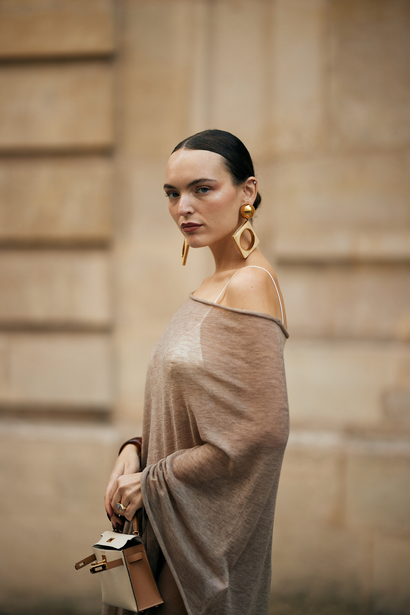 a woman with a brunette bun wearing large gold earrings and an off-the-shoulder tan top