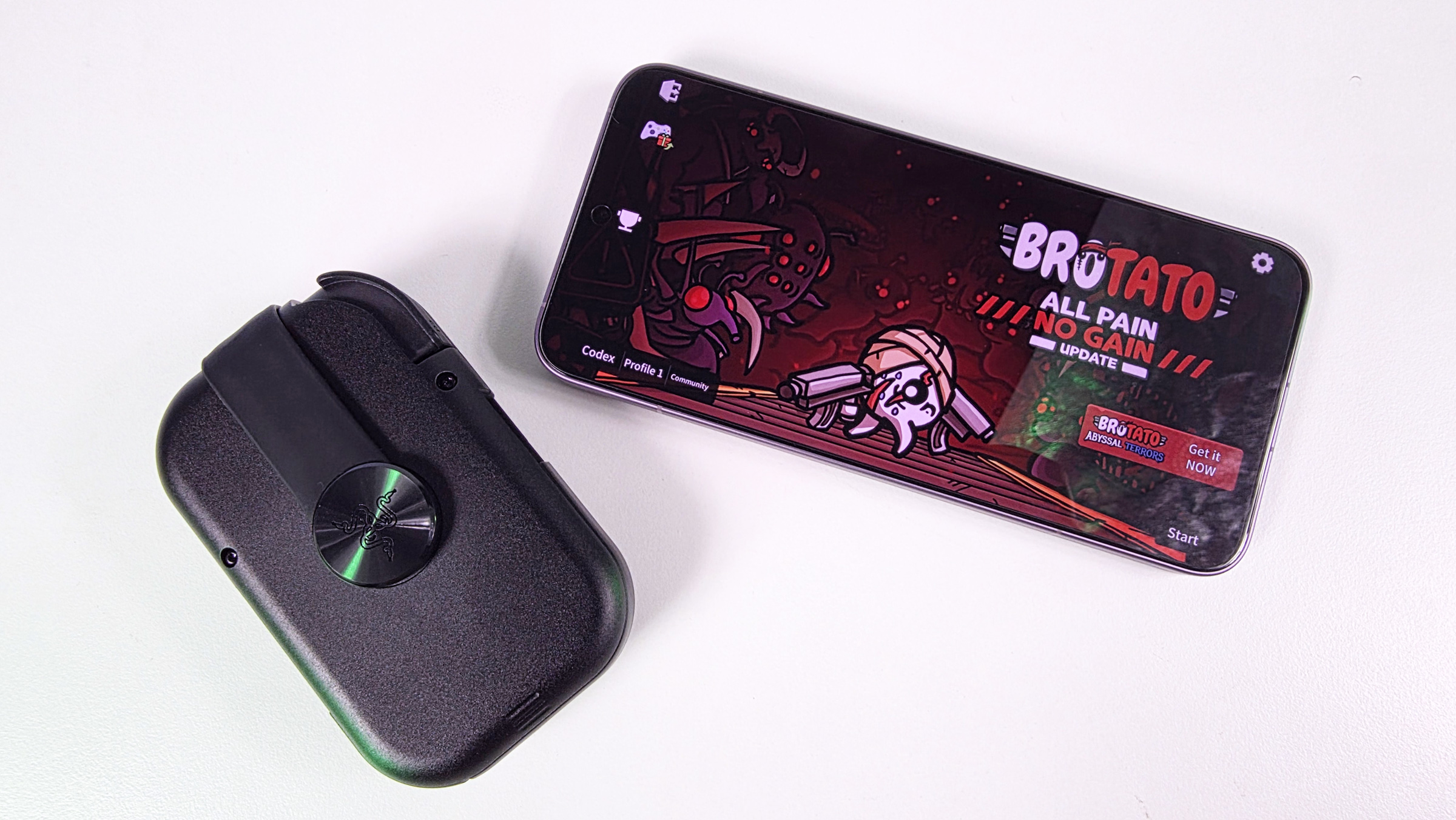Folded, compact Razer Prio mobile controller resting next to a smartphone displaying the main menu of the game Brotato on a white surface.