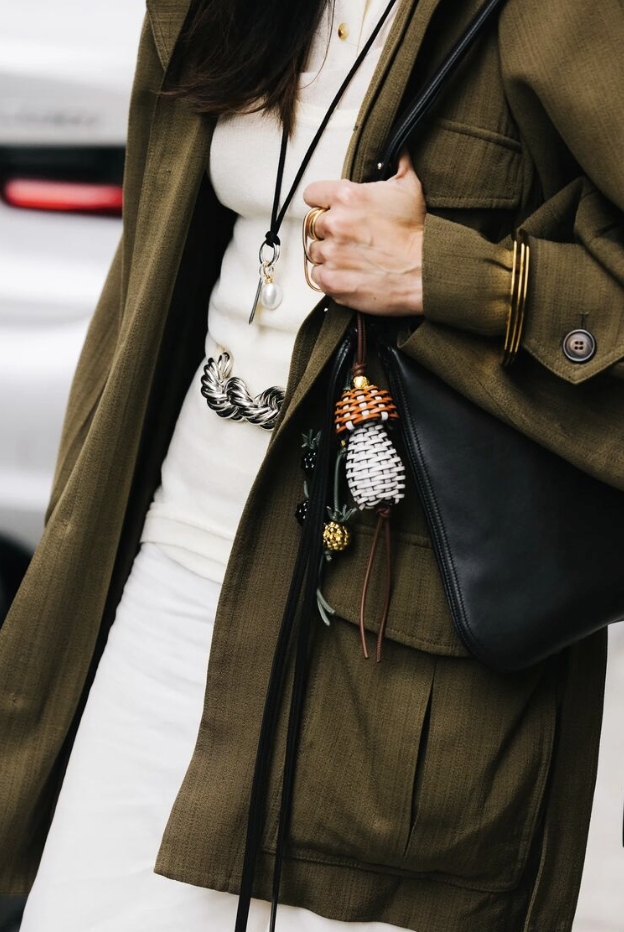 a New York Fashion Week guest wearing a green jacket, white dress, black and pearl pendant necklace, gold bangle bracelets, a black bag with beaded charms, and a silver belt.