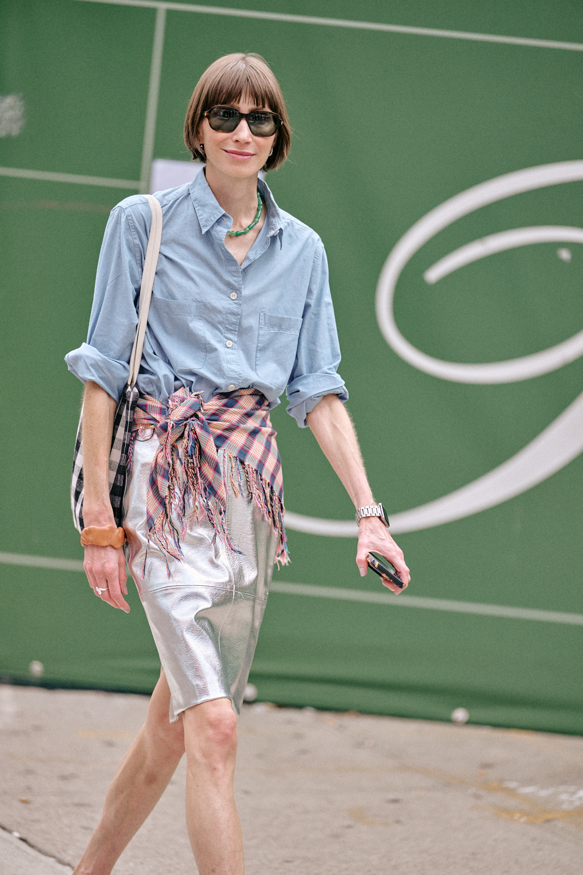 a woman at new york fashion week street style wearing a silver skirt, blue button-down shirt, and plaid pink scarf as a belt