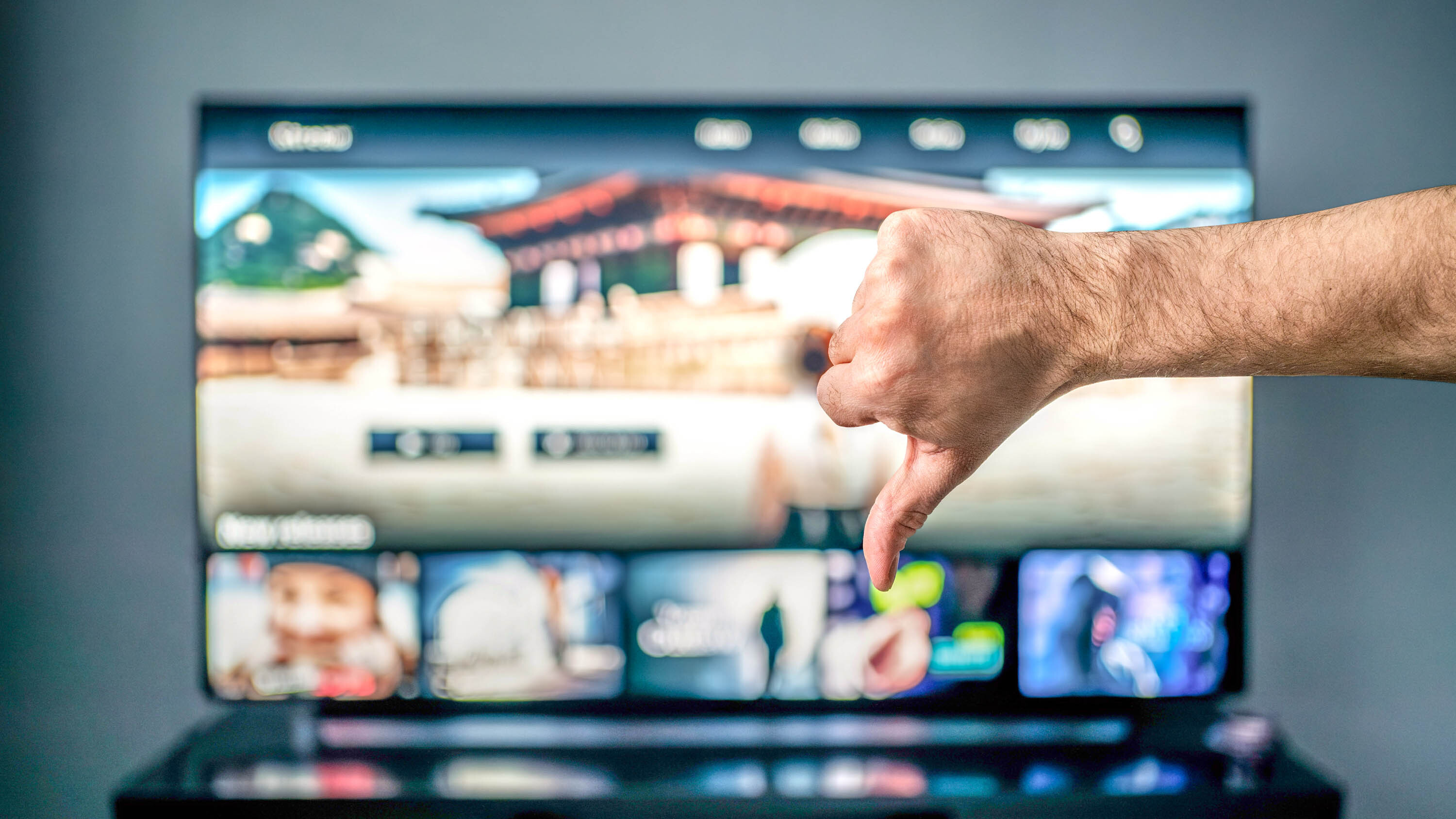 An out-of-focus TV displaying a smart platform home screen is foregrounded by a someone's disembodied "thumbs down" gesture