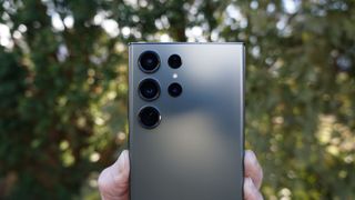 En s&oslash;lvaktig mobiltelefon av typen Samsung Galaxy S23 Ultra holdes i en h&aring;nd, med baksiden vendt mot kamera.