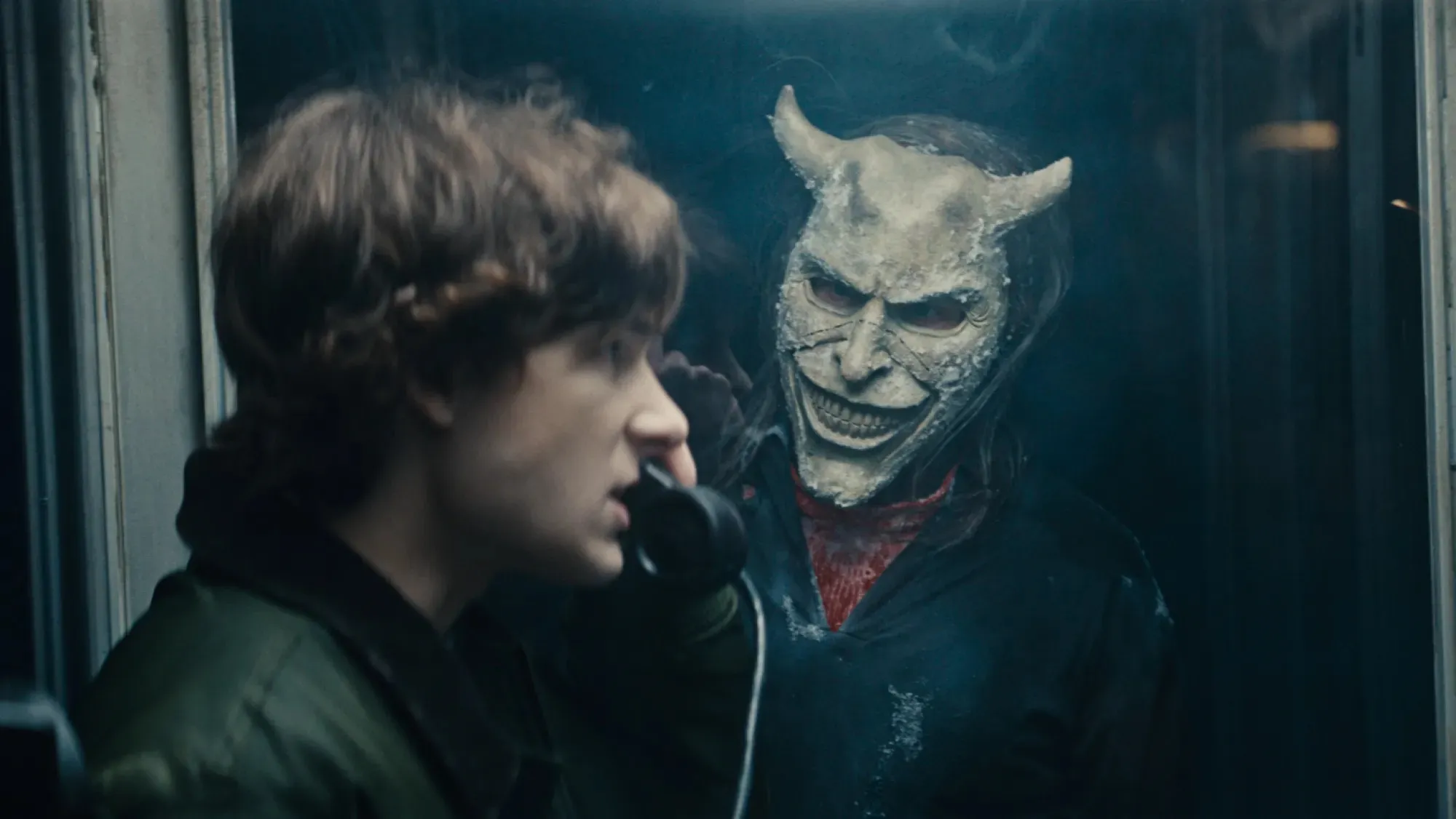 a boy on the phone in a phone booth and a man approaching him in a still from black phone 2