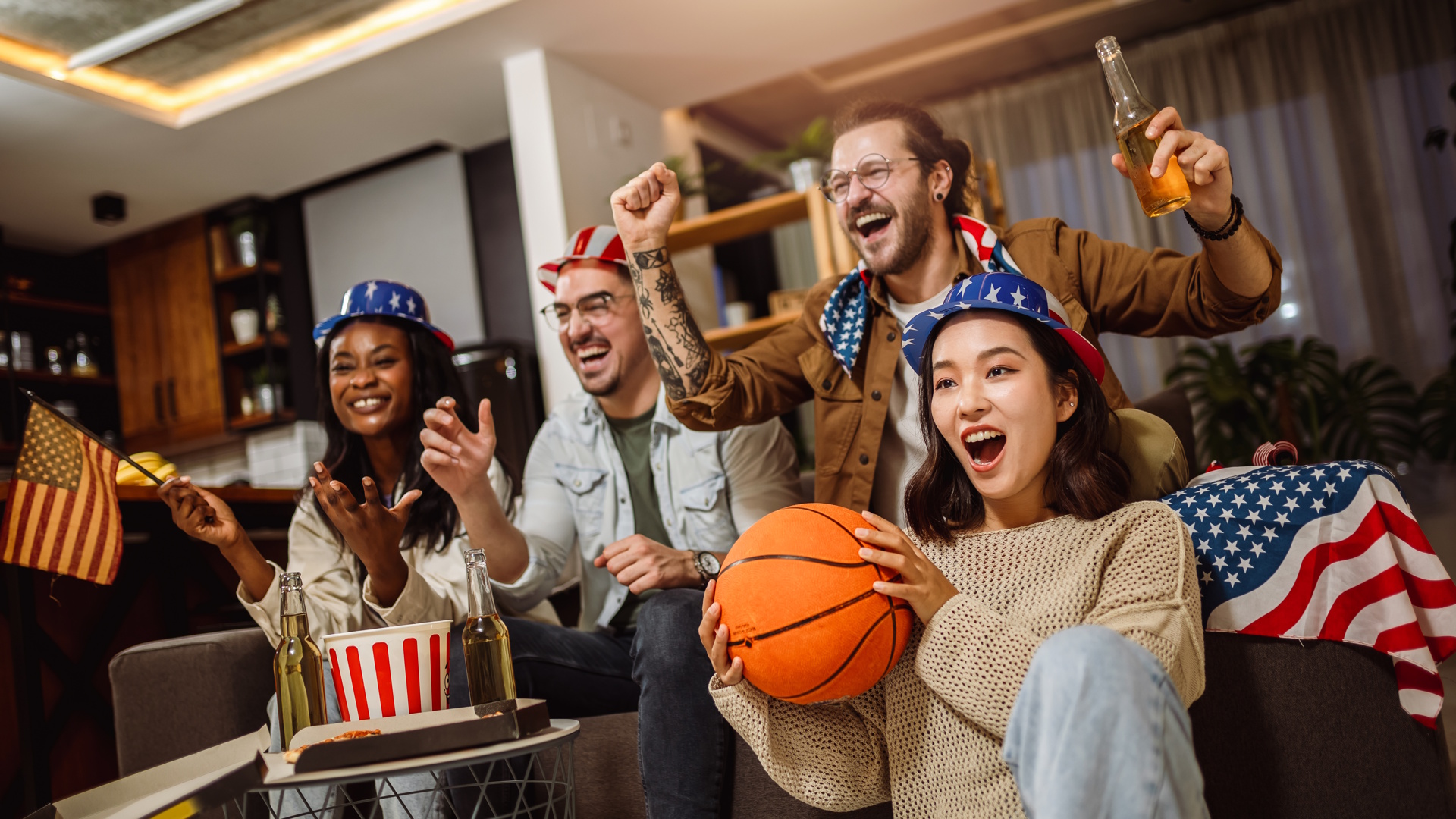 Multiracial group of friends watching basketball game with American flags, drinking beer and cheering.