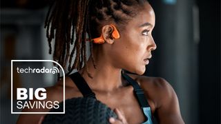 Lifestyle image showing woman working out with medicine ball while wearing Shokz OpenRun Pro 2 headphones in orange color option. A TechRadar 'Big Savings' badge appears overlaid at the bottom left.