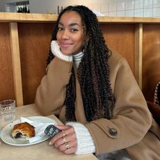 Dutch female fashion influencer Amaka Hamelijnck poses at a cafe wearing a winter outfit with camel coat layered over a chunky white sweater