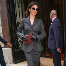 Kendall Jenner wears a gray blazer, gray skirt, burgundy clutch, black leather gloves, and black sunglasses.