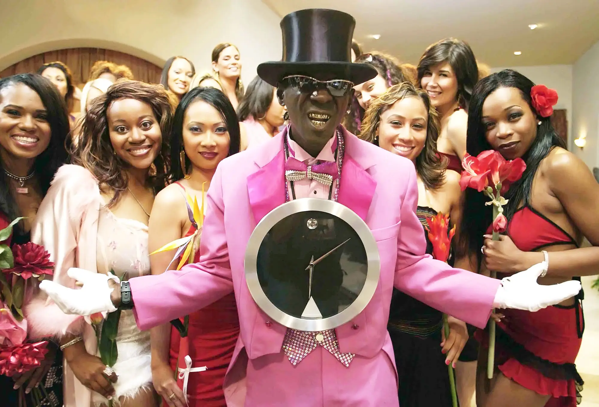 flavor flav poses in a pink suit with a clock around his next surrounded by women in a shot from flavor of love