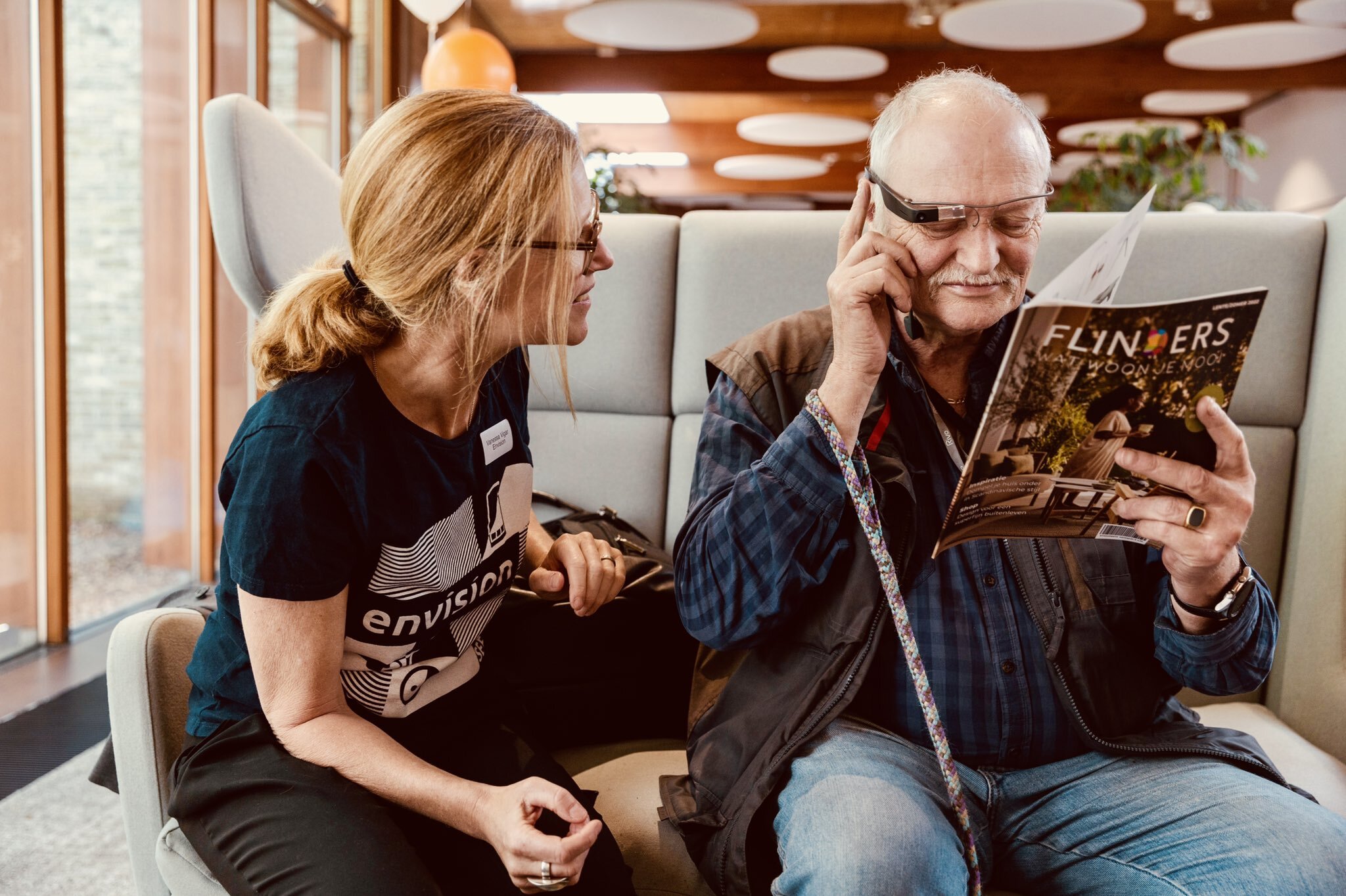 A person trying the Envision Glasses to read a text while another person next to them points the Envision Glasses' camera to a magazine.