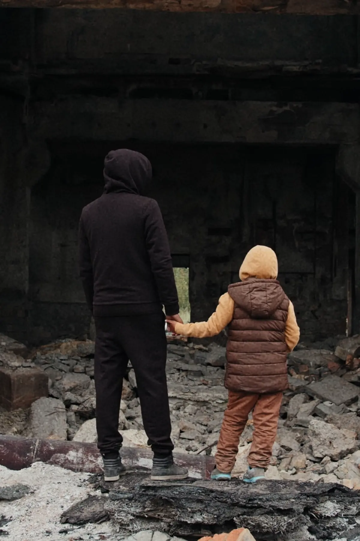 Image of a child holding the hand of a younger child in front of rubble that looks like a destroyed home.