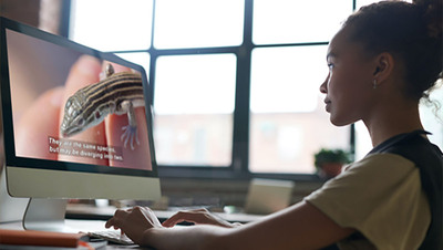 A young girl with her hands on a computer keyboard watches a fullscreen video of a lizard with closed captioning