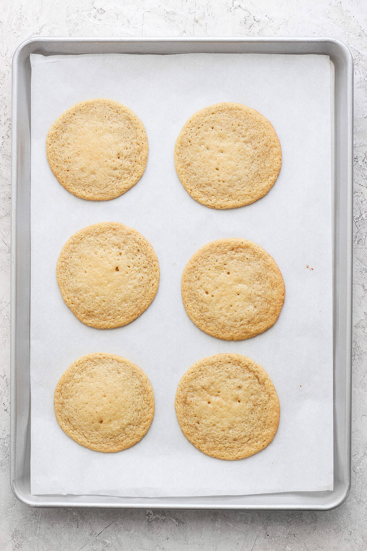 Six round lemon cookies with cream cheese frosting rest on a parchment-lined baking sheet.