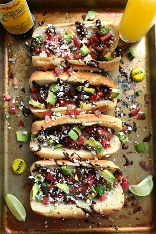 Overhead vertical image of four hot dogs with chopped avocado, crumbled cheese, and a drizzle of mole sauce, with a glass and a bottle of beer, two bottle caps, and lime wedges on a metal baking sheet.