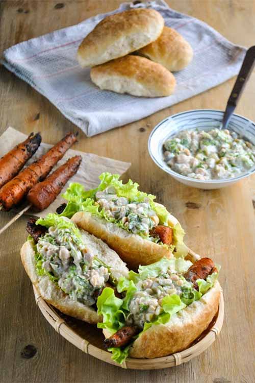 Barbecue carrot dogs with lettuce and chickpea salad, in a basket in the foreground, with three grilled carrots on sticks on a napkin, three hot dog buns on a folded kitchen towel, and a bowl of garbanzo salad with a spoon, on a wood surface.