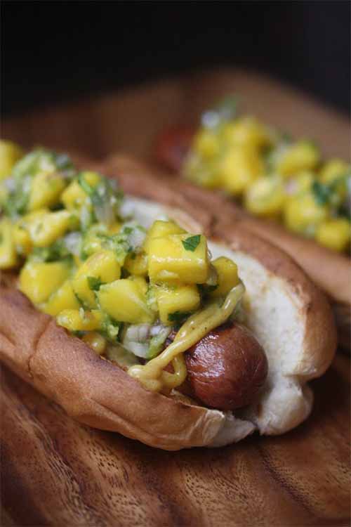 Fresh fruit chutney and Madras curry spiced mayo tops two wieners in buns, on a wood surface.