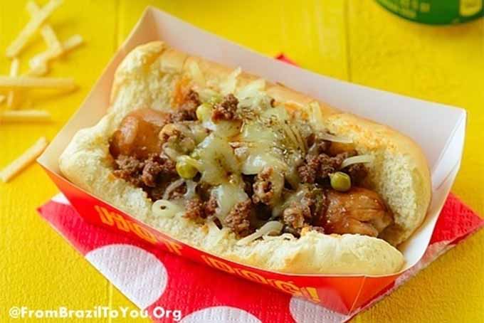 Brazilian Hot Dog topped with melted cheese, in a cardboard holder, on top of a red and white polka-dot paper napkin, on a yellow table with scattered french fries.