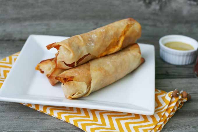 Three Cheese Hot Dog Wontons piled on a square white plate, on top of a yellow and white zig-zag striped folded cloth napkin with a white ramekin of sauce in the background, on a gray wood surface.