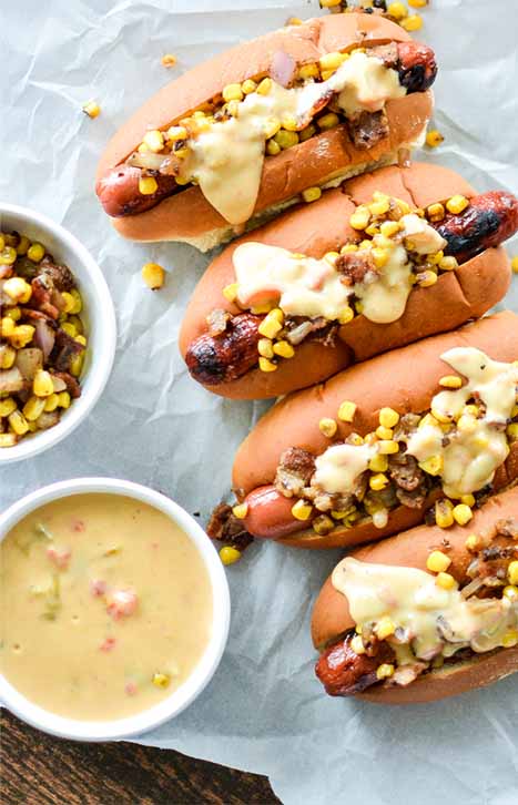 Overhead image of four grilled hot dogs in buns, topped with corn and melted cheese, with two small dishes of cooked corn and queso dip, on a piece of waxed paper on a wood surface.