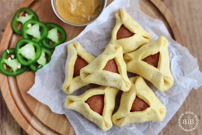 Corn Dog Hamantaschen in a pile on a piece of crumpled waxed paper, with sliced jalapenos and a small cup of mustard, on a round wooden serving platter.