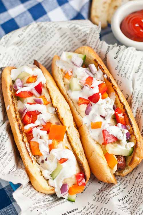 Two grilled franks topped with vegetables and tzatziki, on newspaper on top of a blue and white checkered cloth, next to a small white dish of ketchup.