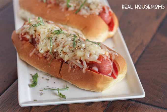 Pizza Hot Dogs topped with melted cheese and fresh herbs, on a white rectangular ceramic serving platter, on a brown wood surface.