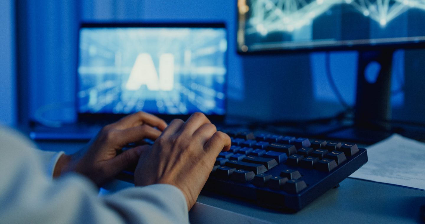 Person types on a keyboard, with a monitor behind showing the letters "AI."