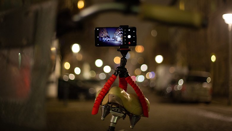 Ein Smartphone, das auf einem Fahrradsattel montiert ist und eine Nachtszene mit verschwommenen Stadtlichtern aufnimmt.