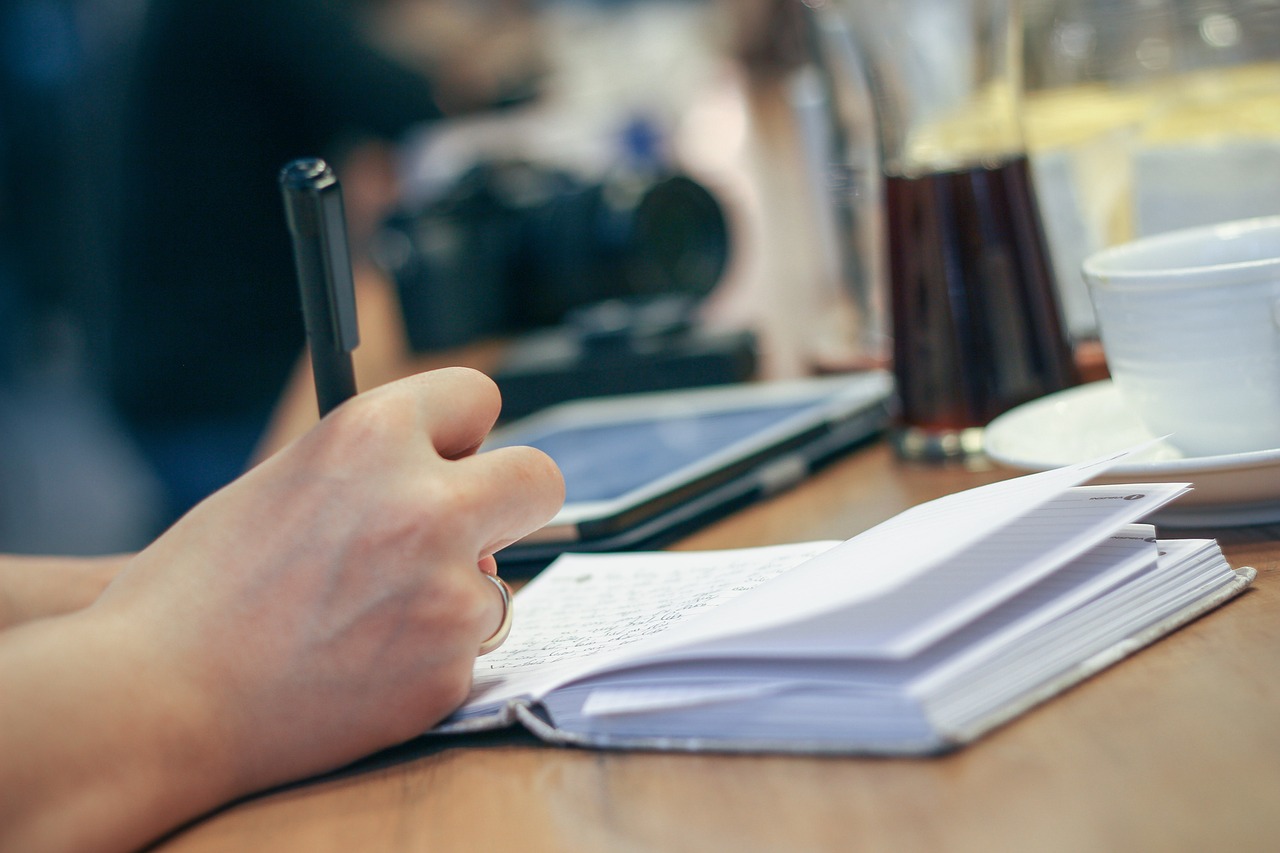 A table with an open notebook sitting upon it, with a hand holding a pen writing in the notebook.
