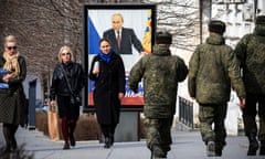 People and soldiers walking past a street poster of Vladimir Putin