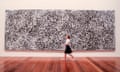 A large painting in black and white, featuring a rhizomatic patterning, hangs on a white gallery wall, with a person walking in front of it.