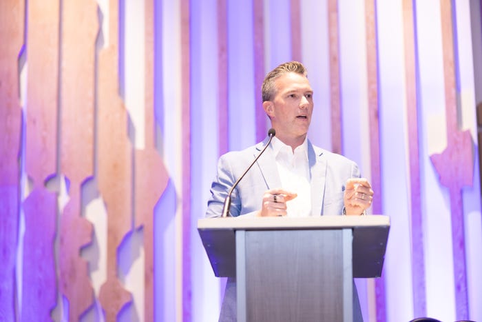 A man stands on stage speaking at a podium with a purple backdrop.