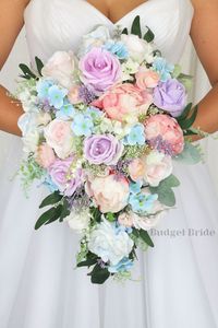 This show-stopping cascading brides’ bouquet is made with a soft and romantic combination of light blue, blush pink and lilac. Light blue accent florals and crystal pins make this bouquet stand out, adding a new twist to the tried-and-true blue rose bouquet. As always, our cascading bouquets are not made using a foam base and are wired by hand, so that you can rest assured your bouquet will maintain both its durability and beauty. This bouquet is approximately 12 inches wide and about 15 inches