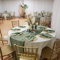 Embrace rustic charm at our event tables. White covers and sage green table runners create a serene base with gold woven placemats and stylishly folded sage green napkins on top. The center hosts candles in themed holders, surrounded by rustic flower arrangements. A Chiara stand backdrop, draped in white and sage green spandex, showcases a boho-themed flower arrangement, completing the rustic allure for an enchanting celebration.