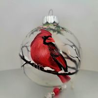 Decorate for the holidays with this cute cardinal ornament.  This ornament displays a single male cardinal. He is sitting on a snowy branch which is painted all around the ornament. The top displays holly leaves and berries to add a pop of green to the ornament. Each ornament is hand painted so each one will be unique. This ornament is painted on a clear globe so the decoration shows through on all sides.  Each ornament will be shipped in a gift box (this may vary depending on availability) and can be personalized before shipping. I have also added a homemade ornament hanger to display your ornament and a note about the symbolization of the red cardinal. The price is per ornament.