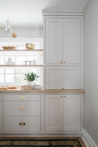 Wood floating shelves are stacked in front of a window, lit by a white and gold sconce, and mounted over and beside light gray cabinets donning vintage brass hardware and a wooden countertop.