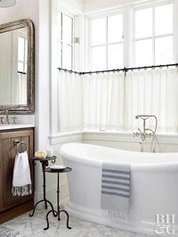 In this tub alcove, multiple windows are treated as one unit. Pleated bathroom window curtains stretch from wall to wall, accenting the window grids. The simple, semi-sheer curtains add an element of privacy, while iron rings add farmhouse style to the look.