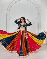 This festive season with this vibrant multi-hued lehenga featuring a stunning mix of colors like deep blue, mustard, orange, and red. The outfit is enhanced with detailed mirror work and charming tassels, giving it a playful touch perfect for Navratri, weddings, or any traditional celebration. The blouse adds a modern twist with intricate embroidery and shell embellishments, making this ensemble a blend of elegance and boho charm.