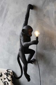 lifestyle image of left-hand hanging monkey wall lamp - black - suitable for outdoors lit up on grey wall with black and white cowhide armchair in background