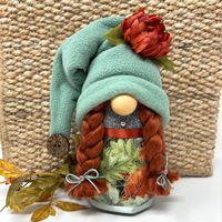 Gnomes can enjoy the fall months when the weather turns cooler because they're well-prepared for the coming winter. That could be why this beautiful decorative female gnome with a fall theme looks so relaxed and cozy. Gnome is approximately 8" (20cm) tall and 4.5" (11cm) around. Made with gray cotton-poly blend fabric stuffed with fill and weighted at the bottom, fleece hat and fabric skirt, and a wooden nose. Her red woolen braids are tied with bows and copper colored flower buttons.  Her hat f