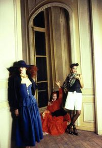  Yves Saint Laurent Rive Gauche Editorial for Vogue. The model on the left poses with her left hand on her hip; she is wearing a blue lace-up cotton poplin bodice with matching skirt and a black cotton poplin jacket with large brimmed black hat, gold necklace and earrings, and black sandals. The model in the middle crouches on the ground with one arm raised above her head; she is wearing a red off-the-shoulder long sleeve chiffon top with a red lace-up cotton poplin bodice and a flounced red skirt with a red headscarf and gold earrings. The model on the right has her right hand on her hip and her left hand up, holding a cigarette; she is wearing a black cotton poplin lace-up bodice with white Bermuda shorts and a floral embroidered silk shawl. Makeup by Jacques Clemente of Elizabeth Arden.